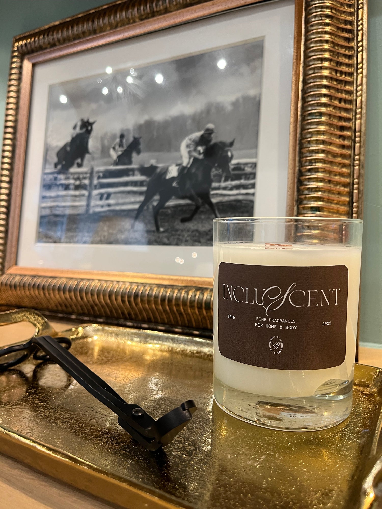 A candle in a glass vessel on a gold plated tray with a black wick trimmer. A picture of a derby is behind in a gold frame. 