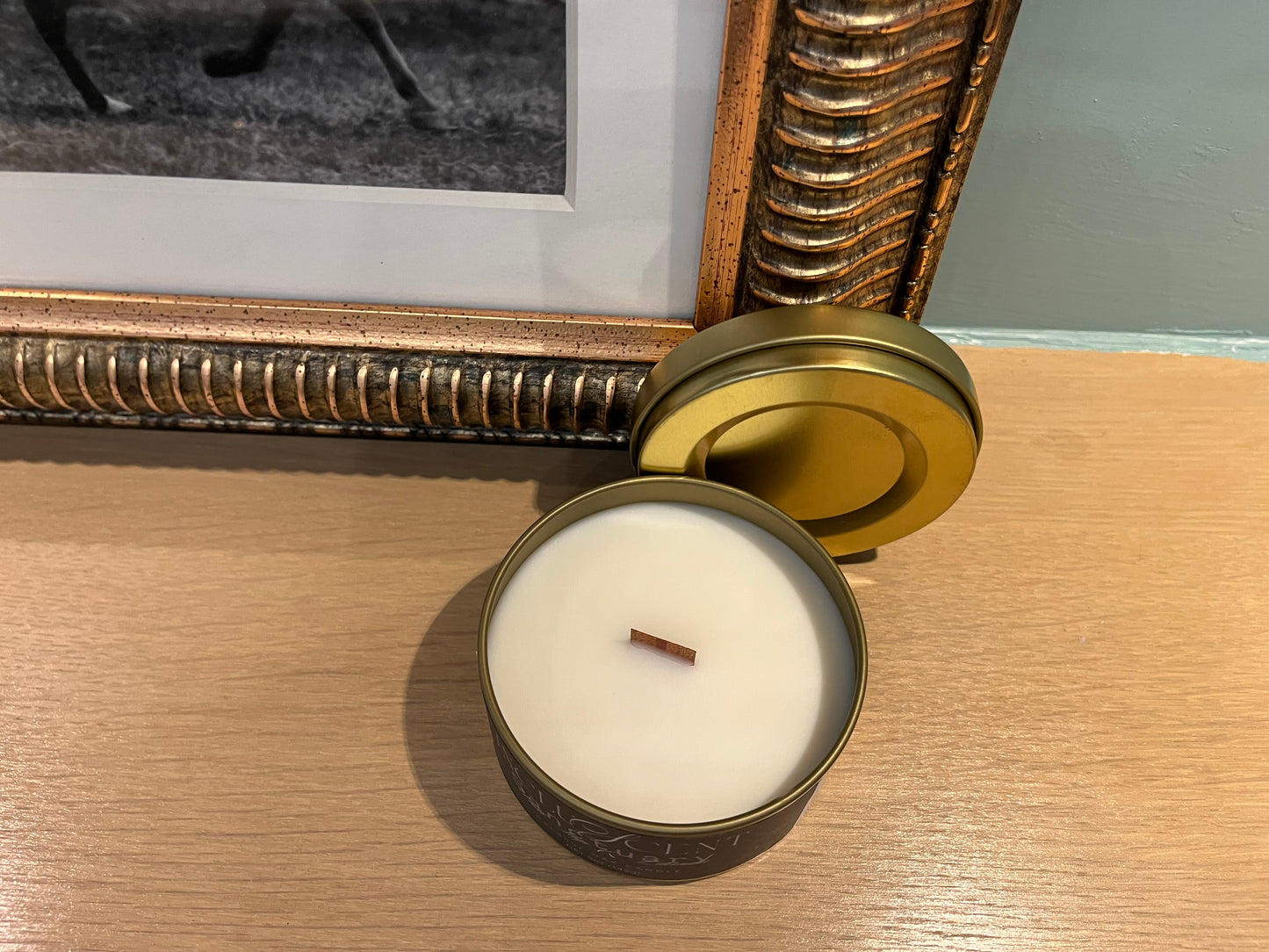 Candle in a metallic tin on a wooden surface with a decorative frame in the background.
