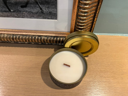 Candle in a metallic tin on a wooden surface with a decorative frame in the background.